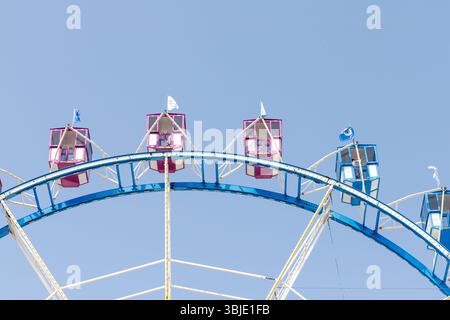 Frammento di una ruota panoramica contro un cielo azzurro, con auto rosa e blu e strutture di supporto bianche. Foto Stock