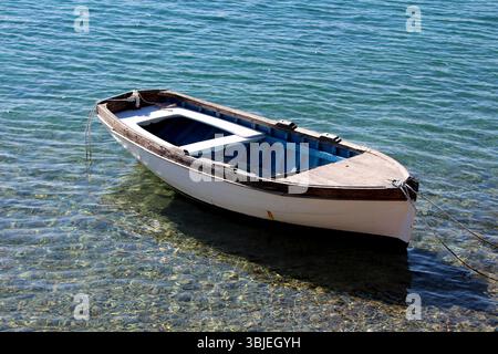 La barca a remi in legno bianca e blu con finiture scure galleggia in acque poco profonde e cristalline, legata da corde a prua e poppa, rivelando ciottoli sottostanti Foto Stock