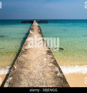 Un lungo molo intempestivo che si estende in un mare turchese calmo, con dolci onde che si infrangono sulla spiaggia sabbiosa. Il cielo è limpido e blu, creando un sereno Foto Stock