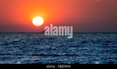 Un tranquillo tramonto sull'oceano, con un grande tramonto all'orizzonte e una piccola barca da pesca che si staglia contro il vibrante cielo arancione. Foto Stock