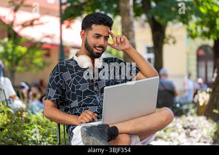 Giovane indiano che usa un computer portatile per celebrare la vittoria di buone notizie sui messaggi, la vittoria del jackpot della lotteria, giveaway online all'aperto. Il giovane turista felice si siede nel parco urbano della strada. Stili di vita della città. Foto Stock