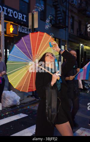 Una giovane donna soffia bolle mentre tiene in mano un ombrello arcobaleno in un evento di strada. Indossa un cappotto nero e abiti scuri. Un'altra persona nella B. Foto Stock