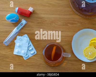 Vista dall'alto della tazza di tè con limone, pillole blister, termometro e inalatore sul tavolo di legno, simboleggiando il trattamento freddo e l'assistenza sanitaria domestica Foto Stock