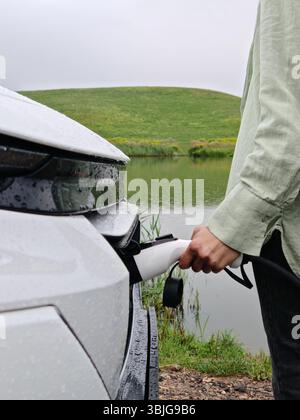 Primo piano di una donna che ricarica un veicolo elettrico utilizzando un cavo di ricarica in un paesaggio rurale, la Bielorussia Foto Stock
