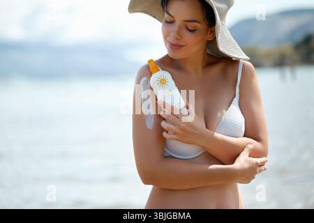 Una bella donna sta su una spiaggia soleggiata, applicando la crema solare per proteggere la sua pelle. Sorride mentre si gode la calda luce del sole e l'atmosfera rilassante Foto Stock