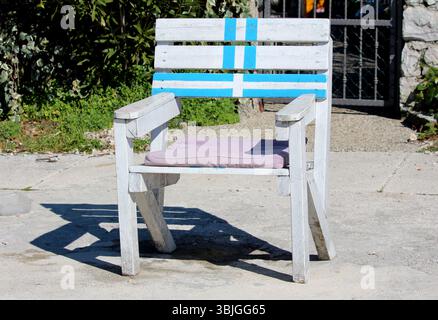 Una robusta poltrona in legno bianco con strisce dipinte di blu e un cuscino di seduta in lavanda si trova su un patio in cemento illuminato dal sole, che getta un'ombra audace Foto Stock