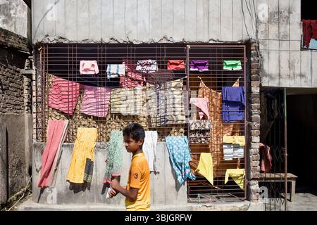 La lavanderia è appesa alle griglie di ferro in un vicolo residenziale a Rajshahi, Bangladesh, mentre un ragazzo passa sotto il sole di mezzogiorno. Foto Stock