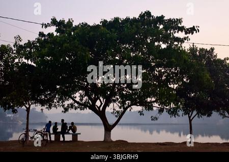 I giovani si riuniscono sotto un albero ombra lungo le rive del lago Kali Ganga al crepuscolo, vicino al Mausoleo di Lalon Shah a Kushtia, Bangladesh. Foto Stock