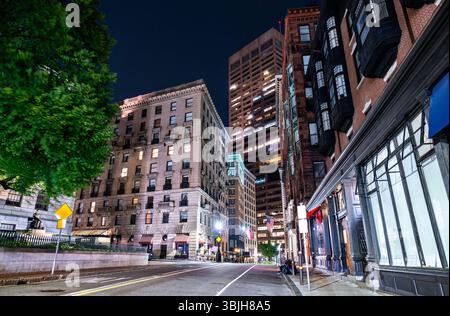 La scena notturna a Boston, Massachusetts, Stati Uniti, mostra una tranquilla strada del centro fiancheggiata da edifici storici in mattoni e un alto e moderno fogliame di alberi che si illumina sotto le luci delle strade e il cielo scuro. Foto Stock