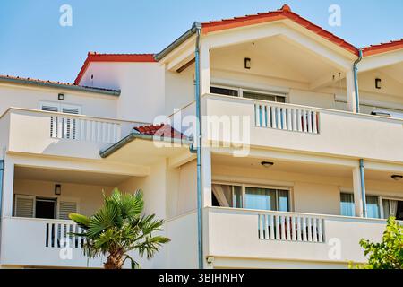 Edificio residenziale bianco con balconi, tetto piastrellato rosso e finestre aperte sotto il cielo blu. Facciata di una villa moderna con terrazze aperte e palme. Io Foto Stock