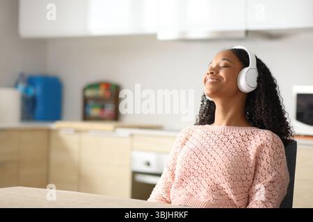 Donna nera felice che indossa le cuffie per ascoltare e rilassarsi in una cucina di casa Foto Stock