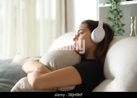 Donna apatica che guarda lontano abbracciando il cuscino che ascolta l'audio seduto sul divano di casa Foto Stock