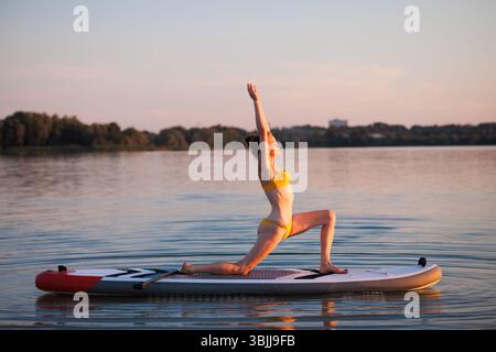 Donna in forma che fa yoga sul paddleboard all'alba, tenendo in posa il guerriero con equilibrio e grazia, circondata da acqua tranquilla e luce naturale soffusa. Foto Stock