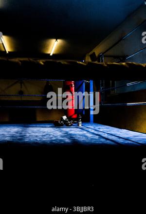 Un anello di boxe si trova in una stanza poco illuminata, illuminata da luci fluorescenti. I guanti da boxe giacciono sul pavimento opaco blu, accanto a una bottiglia di Foto Stock