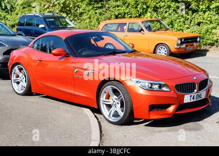 Auto sportiva BMW Z4 metallizzata arancione parcheggiata in un parcheggio soleggiato con auto di scorta ford d'epoca arancione sullo sfondo Foto Stock