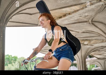 Giovane musicista sorridente e in bicicletta mentre porta la chitarra in una borsa da concerto Foto Stock