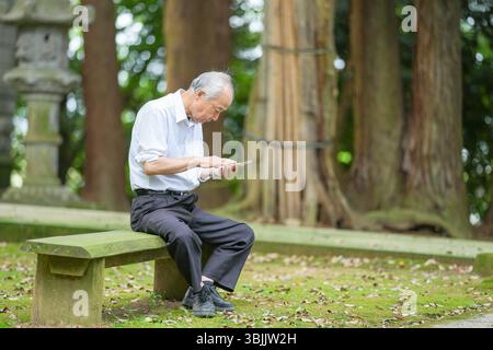 Nel Giappone rurale, un uomo giapponese alla fine degli anni '70 siede in un tranquillo santuario di campagna, indossando una camicia bianca e un cappello. Lui usa il suo smartphone, surrogato Foto Stock
