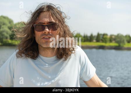 Ritratto di un giovane bruna caucasico con lunghi capelli ricci in occhiali da sole in piedi vicino al fiume. Atmosfera tranquilla e contemplativa. Slo Foto Stock