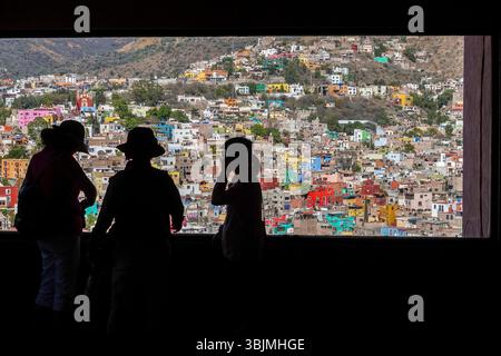 I turisti guardano lo skyline degli edifici color pastello della capitale dalla stazione ferroviaria della funicolare in cima alla collina di San Miguel nella città di Guanajuato, Guanajuato, Messico. La città, patrimonio dell'umanità dell'UNESCO, è costruita in una stretta valle con scalinate sui lati delle montagne ed è conosciuta per la sua affascinante architettura coloniale spagnola. Foto Stock