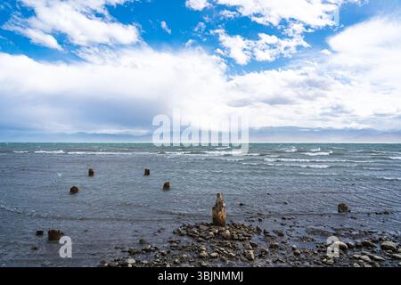 Balykchy, Kirghizistan - 12 maggio 2024: Un ampio colpo di un lago ondulato con pali di legno nell'acqua e un primo piano roccioso sotto un drammatico cielo nuvoloso e. Foto Stock