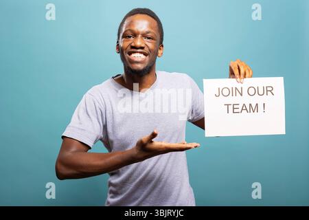 Reclutatore entusiasta che guarda la fotocamera, tiene un documento e invita i candidati a unirsi al suo team. Sorridente afro-americano che punta al segno di assunzione, promuovendo le opportunità di lavoro in un contesto isolato. Foto Stock