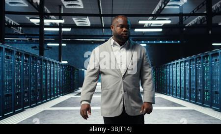 Dipendente che cammina nel data center con spie lampeggianti sulle piattaforme che mostrano l'attività del sistema. Professionista IT che supervisiona server farm scarsamente illuminati con apparecchiature disposte per un funzionamento regolare, telecamera A. Foto Stock