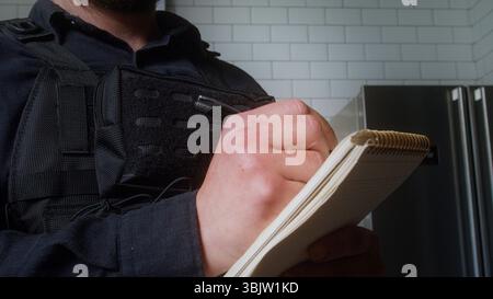 Primo piano di un agente di polizia in uniforme che scrive in un blocco note, forse documentando un rapporto o conducendo un'indagine in casa. Foto Stock
