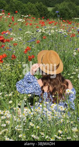 Una donna gioiosa siede in un lussureggiante campo di fiori selvatici adornato da fiori rossi e blu, indossando un cappello di paglia mentre si crogiola al caldo sole. E' un briciolo Foto Stock
