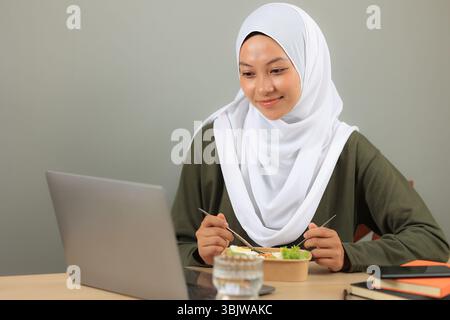 Una bella donna musulmana asiatica che indossa un hijab ama mangiare sano Yummy Salad Bowl mentre usa il suo laptop. Lavoro da casa, vita domestica Foto Stock