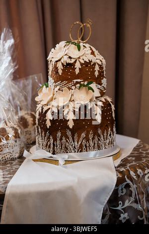 Torta nuziale a due piani splendidamente decorata, decorata con fiori bianchi e frumento, condita da fedi nuziali dorate, perfetta per eventi celebrativi Foto Stock