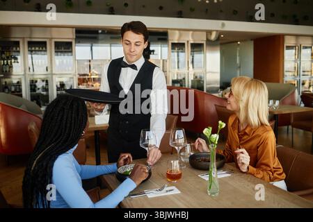 Giovane uomo caucasico che serve cibo per diverse giovani donne nel moderno ristorante Foto Stock