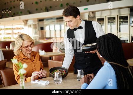Giovane uomo caucasico che serve cibo a donne caucasiche e nere di mezza età Foto Stock