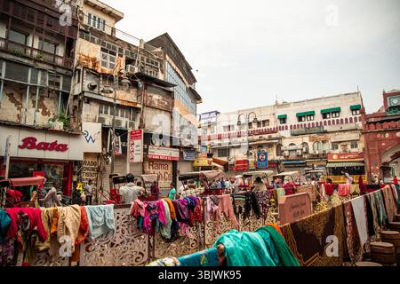 Vestiti colorati appesi su una recinzione in primo piano, risciò in attesa dei clienti e persone sfocate che camminano sullo sfondo nel frenetico mercato Foto Stock