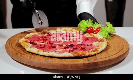 Chef che taglia la pizza Pepperoni. Lo chef affetta i pezzi di pizza. Una vista ravvicinata di uno chef che taglia una pizza al peperoni appena sfornata su una tavola di legno. Foto Stock