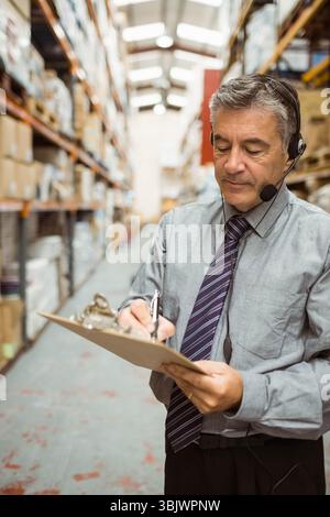 Uomo che supervisiona l'inventario nella corsia del magazzino indossando cravatta, tenendo appunti e cuffie, spazio per le copie Foto Stock