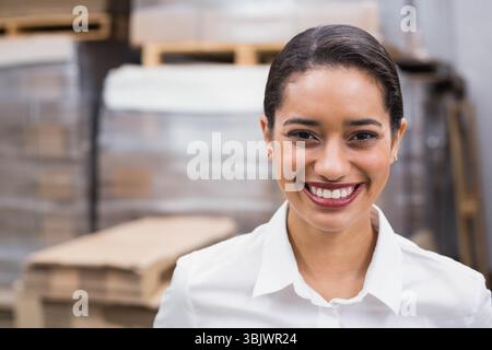 Donna afroamericana adulta sorridente, ispezionando scatole di cartone su pallet vicino agli scaffali del magazzino Foto Stock