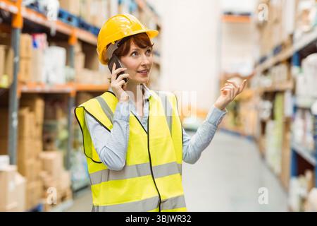 Female manager wearing hard hat and vest gesturing at boxes in warehouse and talking on smartphone Foto Stock