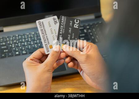Le mani di un uomo in primo piano che usa un computer portatile e tiene in mano carte di credito bianche e nere. Ripresa con vista nera, messa a fuoco selettiva. Tecnologia online pagamento Internet conc Foto Stock