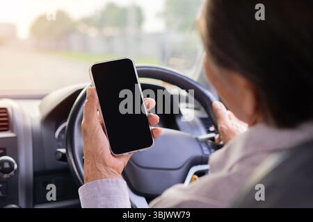 Ripresa della vista posteriore di un uomo asiatico di mezza età che guida un'auto e utilizza uno smartphone con schermo vuoto. Concetto di trasporto, veicolo e tecnologia Foto Stock