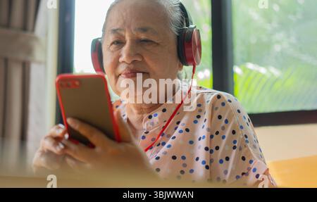 Donna asiatica sicura di sé che tiene in mano uno smartphone mentre ascolta le cuffie in cucina a casa. Concetto di stile di vita pensionistico. Foto Stock