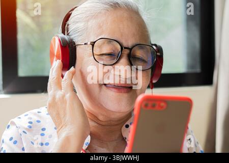 Donna asiatica sorridente e matura con occhiali che ascolta musica con le cuffie e guarda il cellulare a casa. Foto Stock