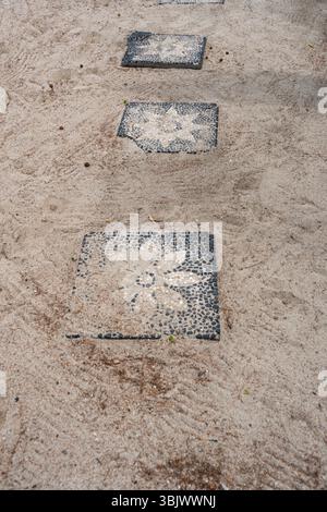 Passerella con piastrelle bianche e nere che hanno dei fiori. La passerella è realizzata in cemento ed è situata su una superficie sabbiosa Foto Stock
