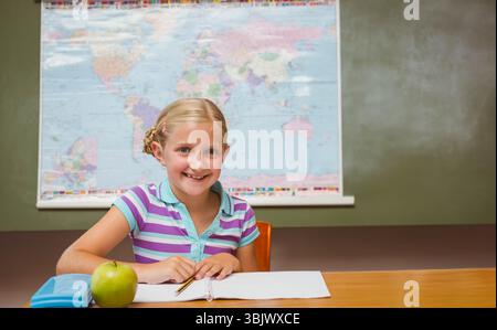Studentessa sorridente mentre si siede alla scrivania della scuola sotto la mappa del mondo scrivendo in un taccuino con una matita Foto Stock