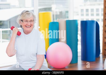 Donna anziana che sorride e solleva il manubrio rosso a spalla sulla panchina nella sala fitness, spazio fotocopie Foto Stock