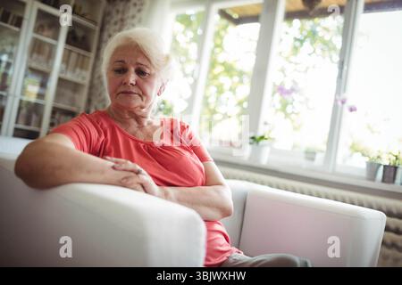 Donna anziana che indossa una camicetta di corallo seduta nella poltrona del soggiorno vicino alla libreria e alle piante in vaso Foto Stock