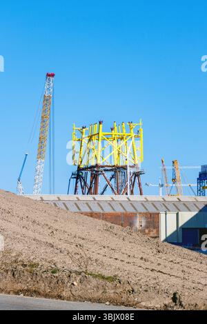 Cantiere eolico offshore con una grande struttura in acciaio giallo, gru e collina di sabbia sotto un cielo azzurro cristallino a lato della spiaggia Foto Stock
