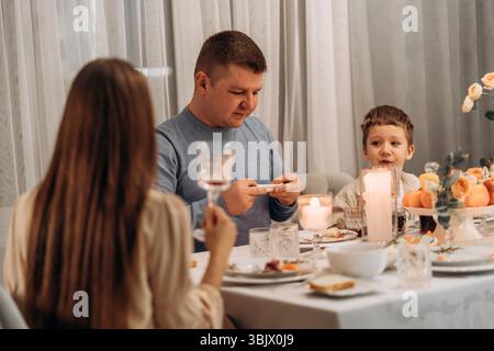 Padre e figlio condividono un'ottima cena a un tavolo accogliente con candele, frutta e fiori, mentre la madre ascolta in primo piano catturare l'autenticità Foto Stock