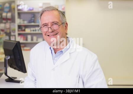 Farmacista di sesso maschile senior in camice da laboratorio in piedi dietro il bancone della farmacia utilizzando il monitor del computer Foto Stock