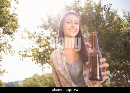 Donna che tiene una bottiglia di vetro marrone verso la macchina fotografica mentre sorride sotto gli alberi del parco illuminati dal sole Foto Stock