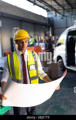 Uomo asiatico che rivede il progetto indossando un elmetto giallo e un giubbotto riflettente nella baia del magazzino Foto Stock
